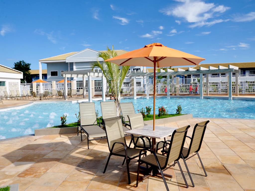 Área da piscina do Lacqua diRoma, em primeiro plano mesa com quatro cadeiras e guarda-sol, logo atrás três espreguiçadeiras e ao fundo a piscina, mais ao fundo prédio de apartamentos.